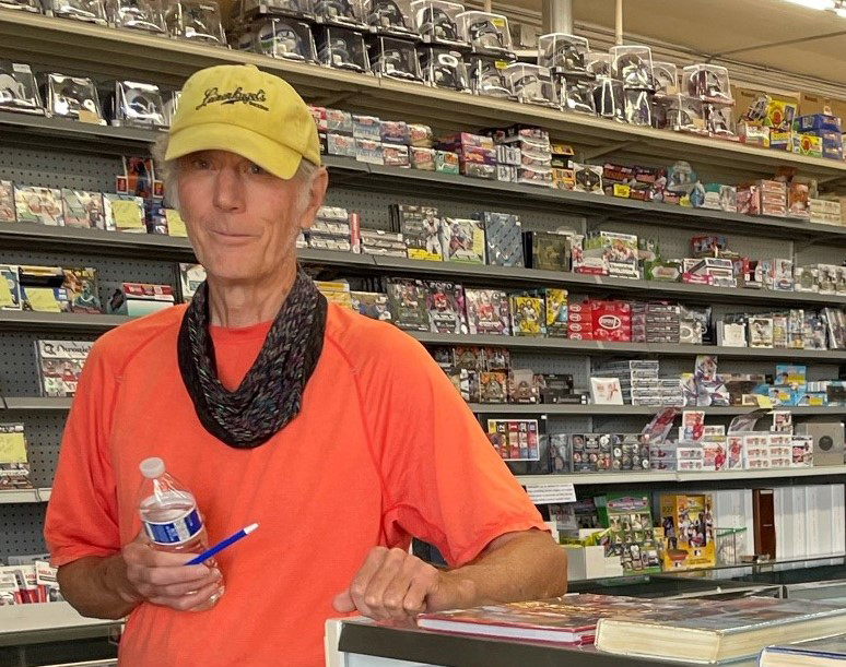 Stan Opdyke wears an orange shirt and stands in a shop surrounded by baseball memorabilia