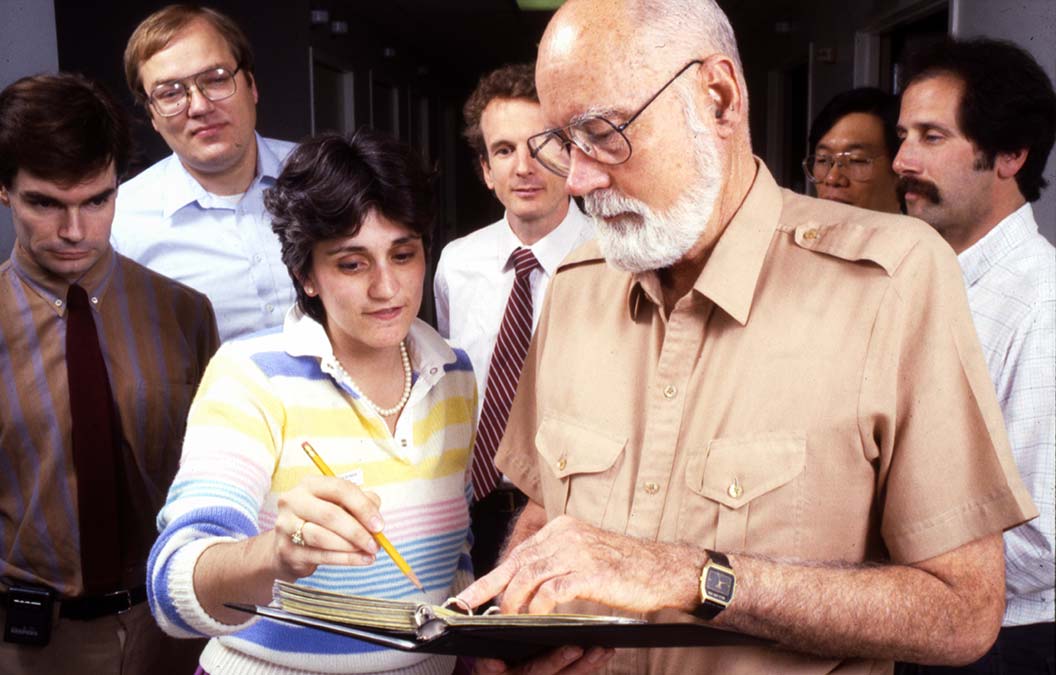 Dr. E. Donnall Thomas with white hair and beard wearing a tan shirt, consulting with a younger woman in a striped sweater who is pointing at notes on a clipboard he's holding. They are surrounded by several men in business attire who appear to be observing.